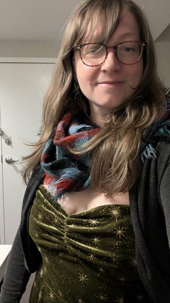 A woman with glasses and blonde hair, wearing a scarf and a green and gold lowish cut romper with a gray sweater and a scarf.